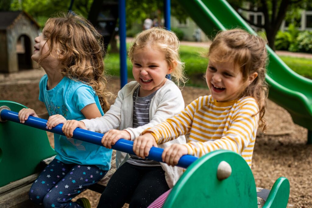 Three young children, two girls with long hair and a girl with shorter hair, are smiling and laughing while sitting together on a green seesaw in a playground. A green slide and a small playhouse are visible in the background.